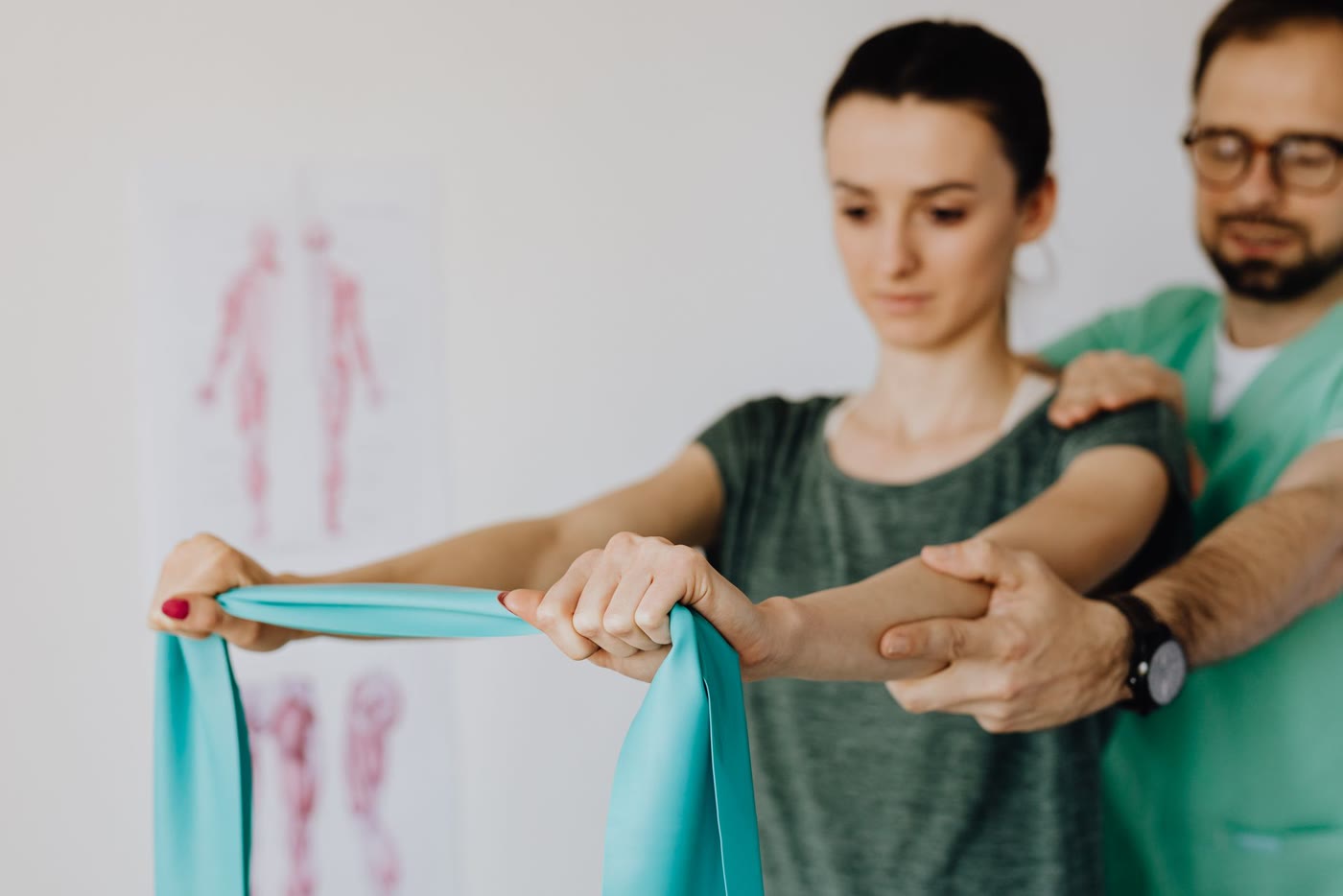 A patient pulls a teal resistance band with both arms while a physiotherapist supports her elbow