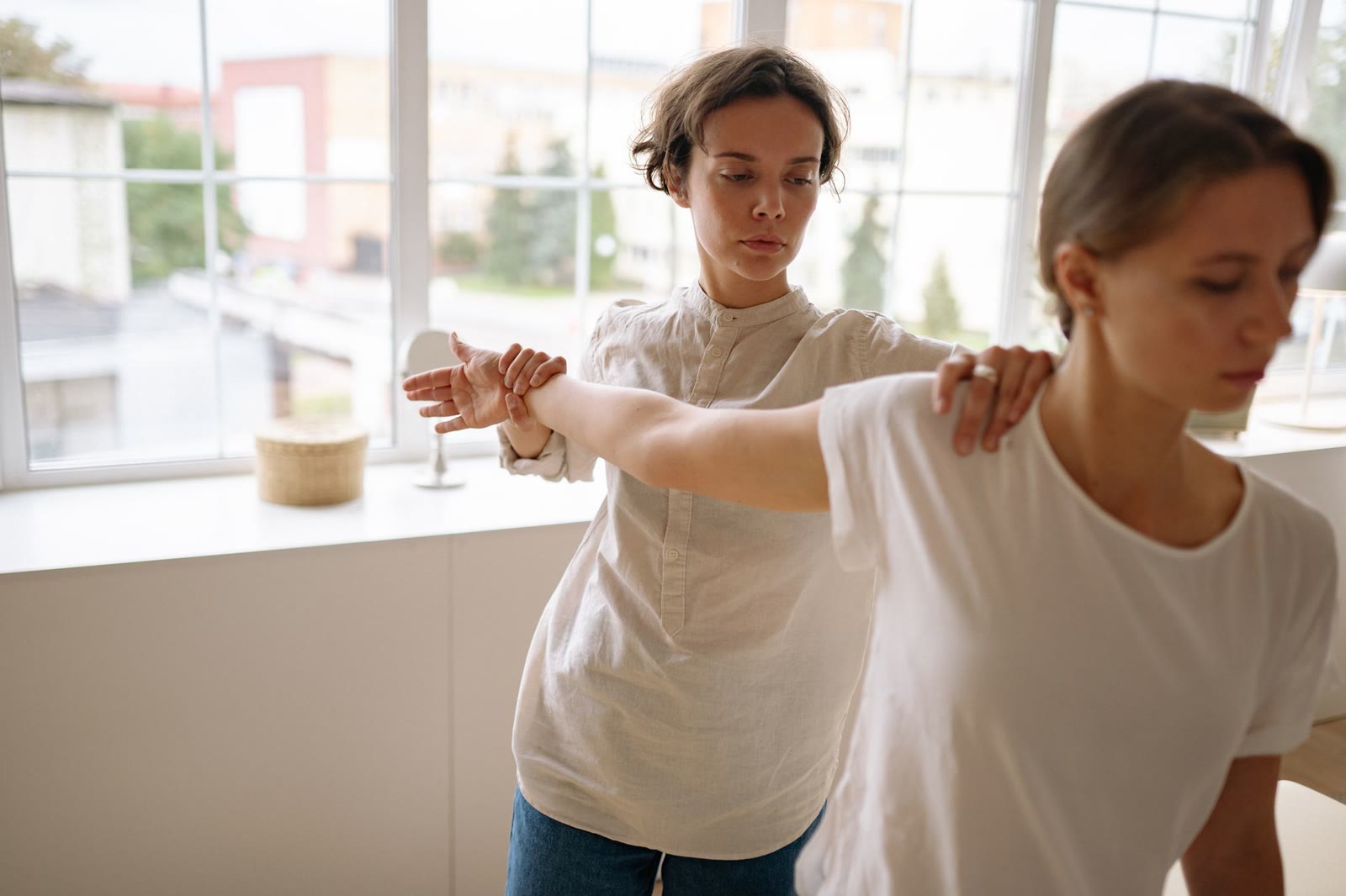 A physiotherapist guides a patient's raised arm through a supported shoulder movement in a bright clinic room